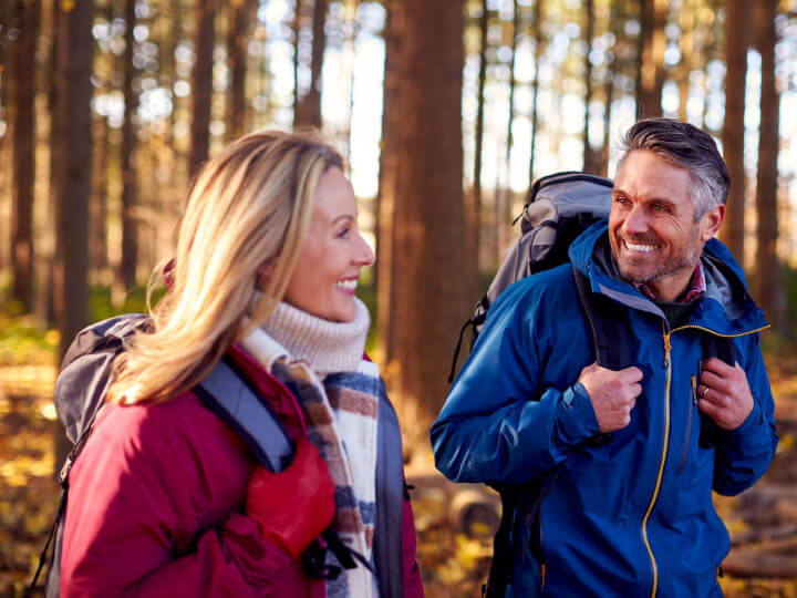 Natural Pharmaceuticals Movisan™ Collagen II Vyriška ir moteriška pora žygiuoja rudens miške su kuprinėmis, dėvėdami šiltus drabužius ir šypsodamiesi.