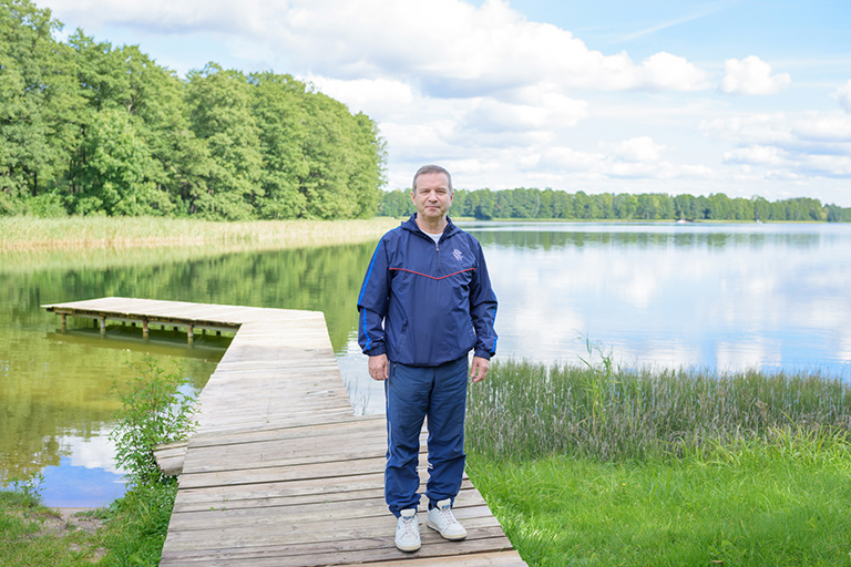 Natural Pharmaceuticals Movisan™ Collagen II Vyriškis vilki sportinį kostiumą, stovintis ant medinio liepto prie ežero su žaliais krūmais ir debesuota dangumi.