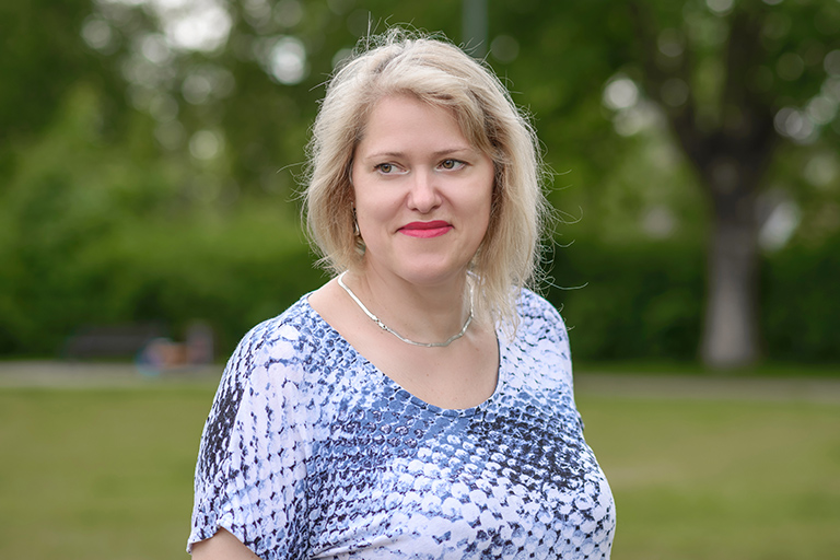 Natural Pharmaceuticals Movisan™ Collagen II Vidutinio amžiaus moteris su šviesiais plaukais, dėvi mėlynos ir baltos spalvų palaidinę, stovi lauke, žiūrėdama į šalį.