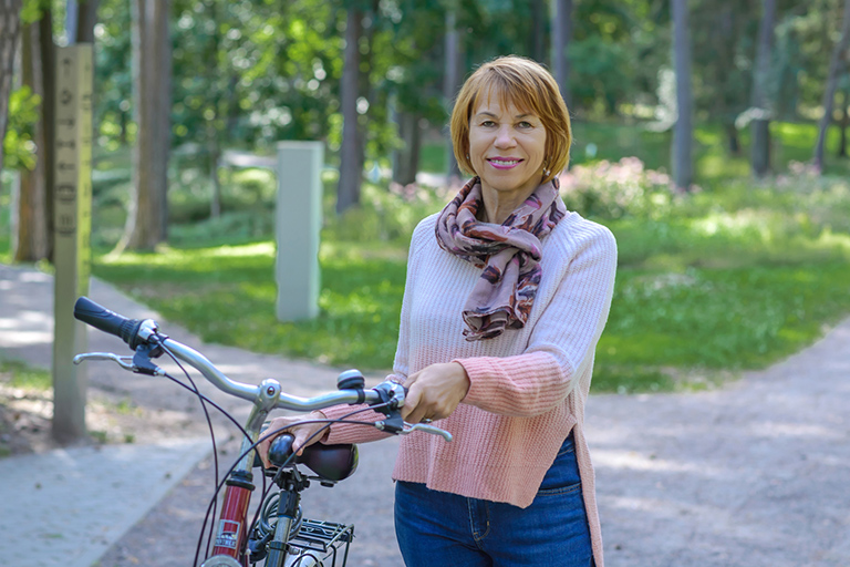 Natural Pharmaceuticals Movisan™ Collagen II Vidutinio amžiaus moteris su dviračiu parke, dėvinti rožinį megztinį ir šaliką.