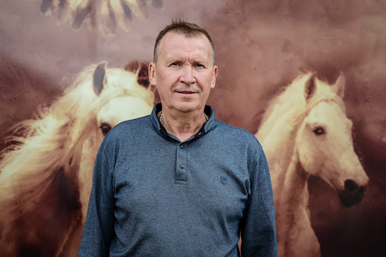 Natural Pharmaceuticals Movisan™ Collagen II Vyriškis mėlyna palaidine stovi priešais fono paveikslą su dviem baltomis arklio figūromis.